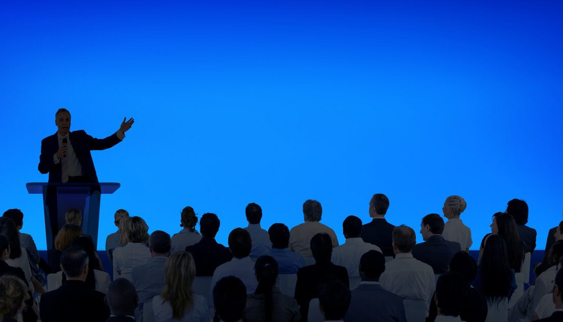Speaker presenting to a large seated audience at a hybrid business event