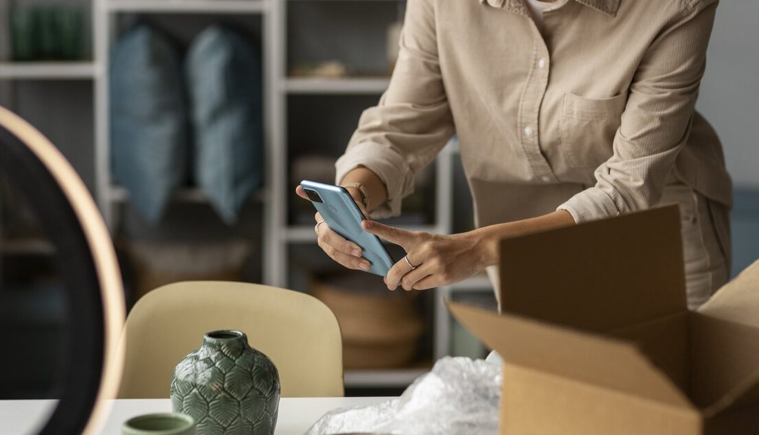 Online seller photographing a product with a smartphone beside a shipping box and ring light