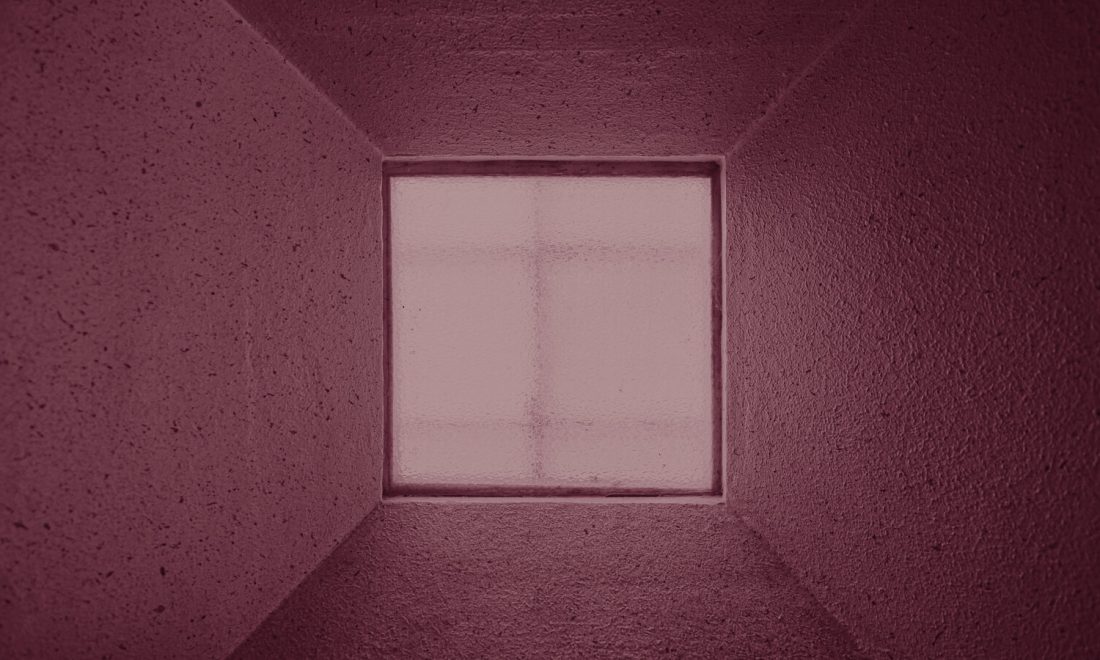 A small square frosted window set deep in a thick concrete wall, admitting light but no detail