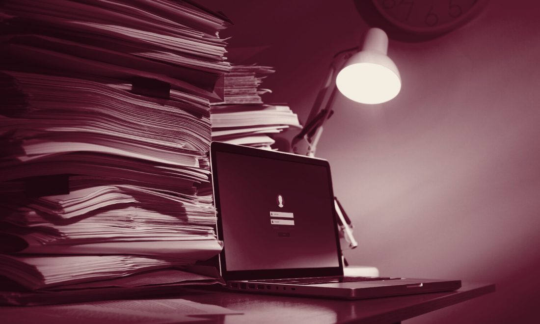 Towering stacks of paper files next to a laptop displaying a locked login screen under a desk lamp