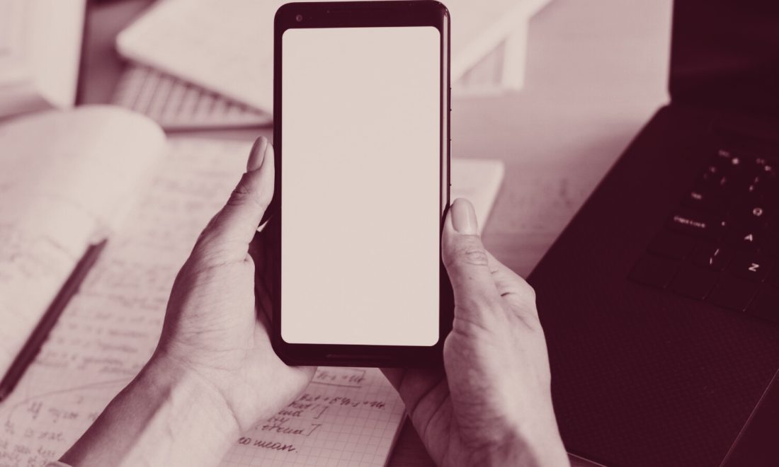 Hands holding a smartphone beside an open laptop on a desk, in wine tones